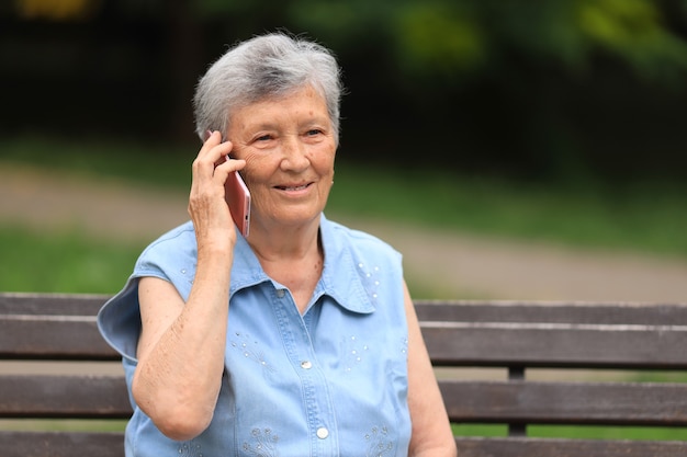 Glückliche ältere behinderte Frau, die auf einer Bank draußen in einem Sommerpark sitzt und am Telefon spricht