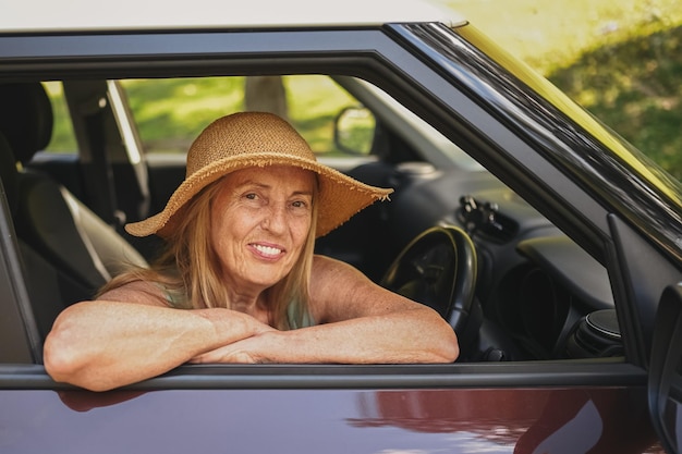 Glückliche ältere Fahrerin mit Strohhut, die im neuen Auto sitzt und lächelnd in die Kamera schaut und genießt