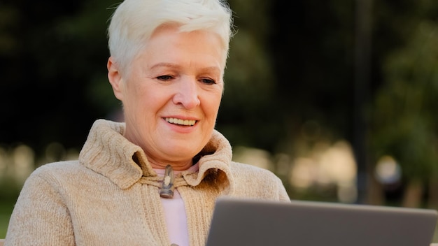 Glückliche ältere Frau, die am Laptop arbeitet, lächelnde ältere Frau im Rentenalter, die Computer durchsucht oder im Internet surft und Nachrichten online liest, aufgeregte ältere Dame, die eine SMS-Nachricht am PC schreibt, die im Freien sitzt