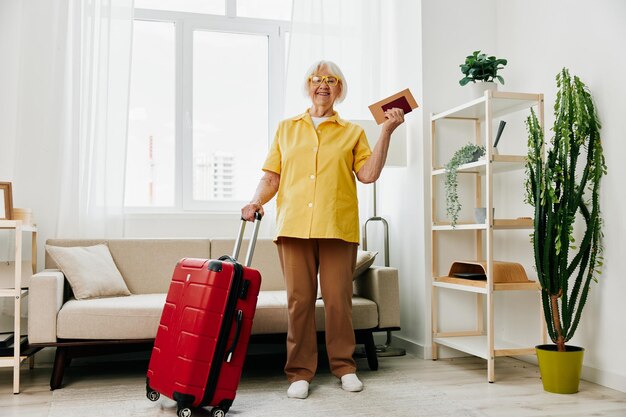 Foto glückliche ältere frau mit reisepass und reiseticket, gepackt in einem roten koffer, urlaub und gesundheitsfürsorge. lächelnde alte frau steht freudig vor der reise im haus