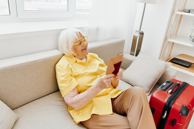 Glückliche ältere Frau mit Reisepass und Reiseticket, gepackt in einen roten Koffer, Urlaub und Gesundheitsfürsorge. Lächelnde alte Frau, die vor der Reise freudig auf dem Sofa saß, hob vor Freude die Hände