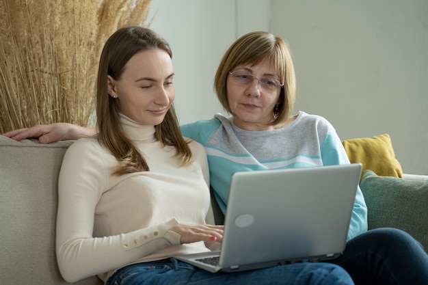 Glückliche ältere mittlere Mutter, die auf Couch mit ihrer Tochter sitzt, die Laptop betrachtet