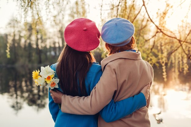 Glückliche ältere Mutter mit Blumen und ihrer erwachsenen Tochter, die am Frühlingsfluss bei Sonnenuntergang umarmt