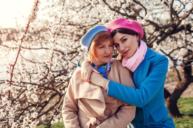 Glückliche ältere Mutter und ihre Tochter, die im blühenden Frühlingsgarten bei Sonnenuntergang umarmen