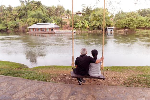 Glückliche ältere Paare, die auf Schwingen zusammen nahe Flusswarteuntergang sitzen.