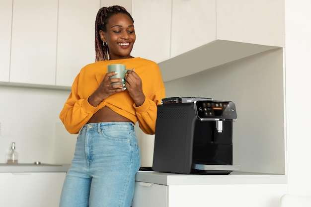 Glückliche afrikanisch-amerikanische Frau, die frischen aromatischen Kaffee in der Nähe einer modernen Maschine im Kücheninterieur genießt