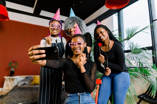 Glückliche afrikanische Mädchen feiern Geburtstag in Hüten und fotografieren auf einem Smartphone