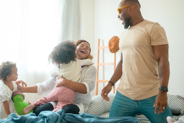 Glückliche afroamerikanische Eltern mit kleinen Töchtern, die zu Hause auf dem Bett im Schlafzimmer spielen