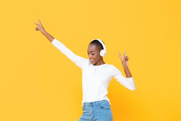 Glückliche afroamerikanische Frau, die Musik auf drahtlosem Kopfhörer mit den Händen oben lokalisiert auf gelber Wand hört