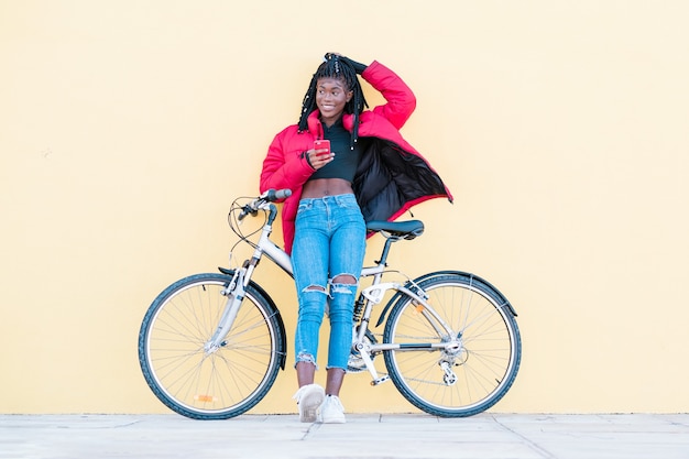 glückliche afroamerikanische Frau mit roter Jacke mit Fahrrad auf der Straße über gelbem Hintergrund