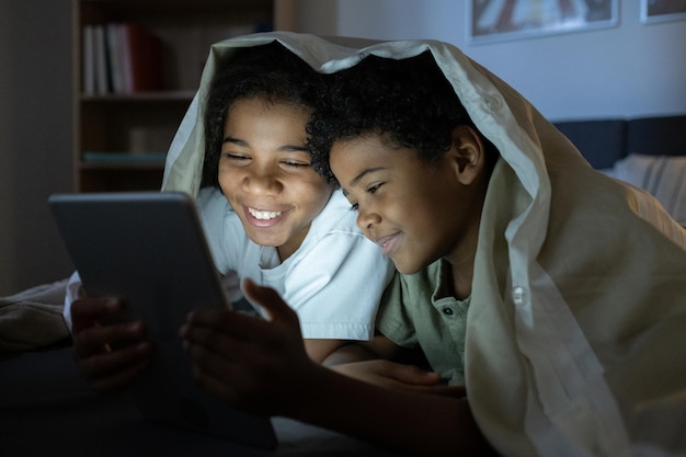 Glückliche afroamerikanische Kinder, die unter der Bettdecke liegen und ein Online-Buch auf einem Tablet im dunklen Raum lesen