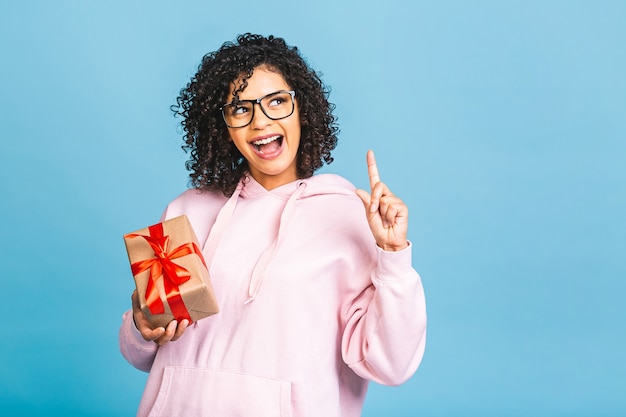 Glückliche afroamerikanische lockige Dame im lässigen Lachen, während Geschenkgeschenkbox lokalisiert über blauem Hintergrund hält.