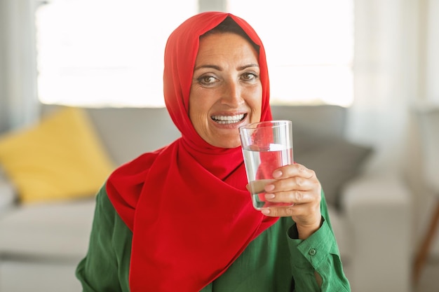 Glückliche alte muslimische Frau trinkt Wasser mit Glas und lächelt in die Kamera, die zu Hause im Wohnzimmer sitzt