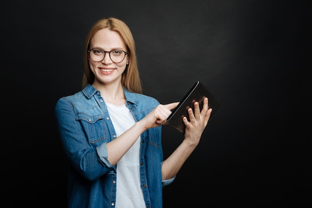 Glückliche amüsierte junge Frau, die Positivität ausdrückt und lächelt, während Tablette benutzt und im Studio steht