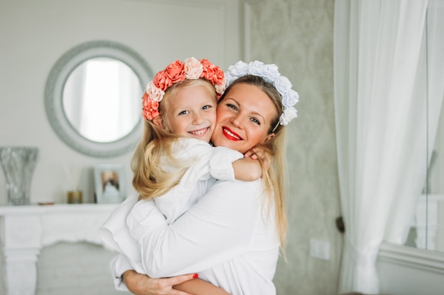 Glückliche angemessene lange Haarmutter und nette Tochter in den Kränzen von Blumen am Wohnzimmer, glücklicher Familienlebensstil