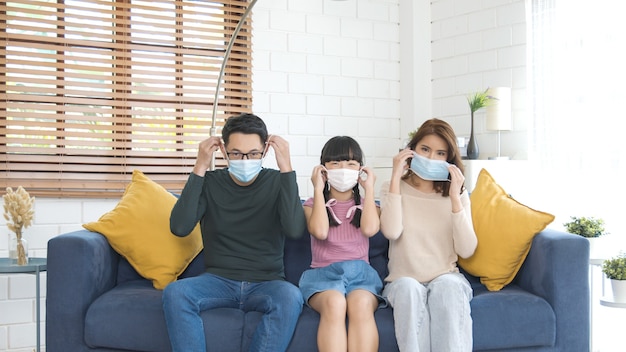 Glückliche asiatische Familie, die eine Maske trägt, um vor Viren im Wohnzimmer zu Hause zu schützen.