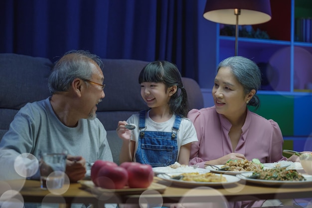 Glückliche asiatische Familie, Großmutter, Großvater und Enkelin, die am Tisch essen und Spaß haben, während zu Hause abends ältere Eltern und Kinder gemeinsam im Wohnzimmer drinnen essen