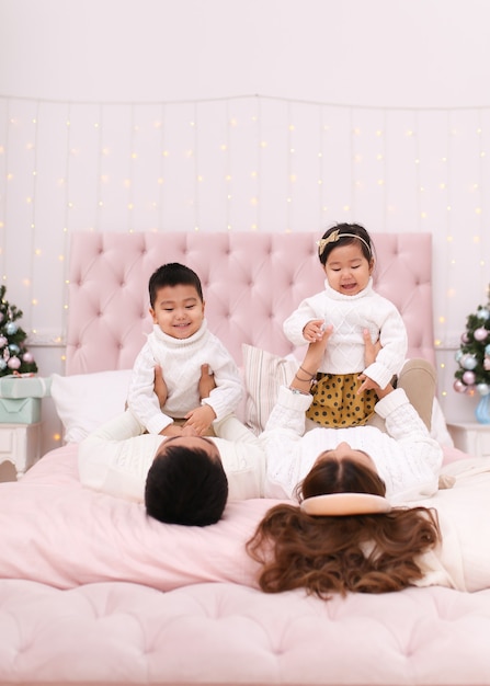 Glückliche asiatische Familie mit zwei Kindern in gemütlichen bequemen Pullovern, die Spaß haben und auf dem Bett entspannen