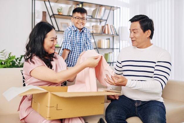 Glückliche asiatische Frau, die hellrosa Kleid aus Geschenkbox von ihrem Ehemann und jugendlichen Sohn nimmt