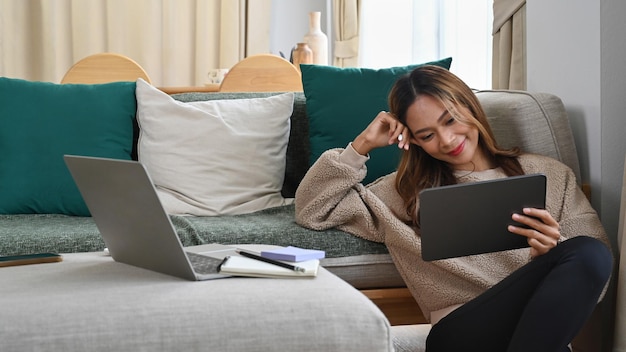 Glückliche asiatische Frau, die im Wohnzimmer auf dem Boden sitzt und mit einem digitalen Tablet im Internet surft