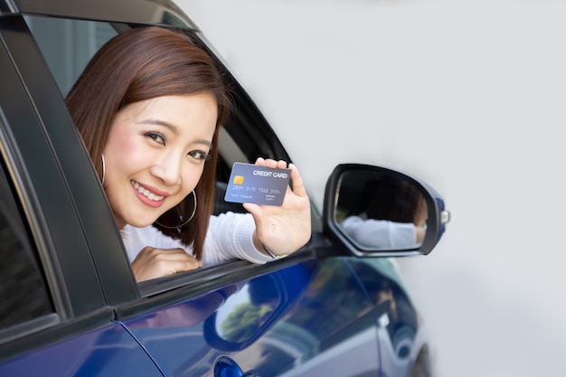 Glückliche asiatische Frau, die Kreditkarte hält, zeigen die Karte und lächeln auf dem Auto.