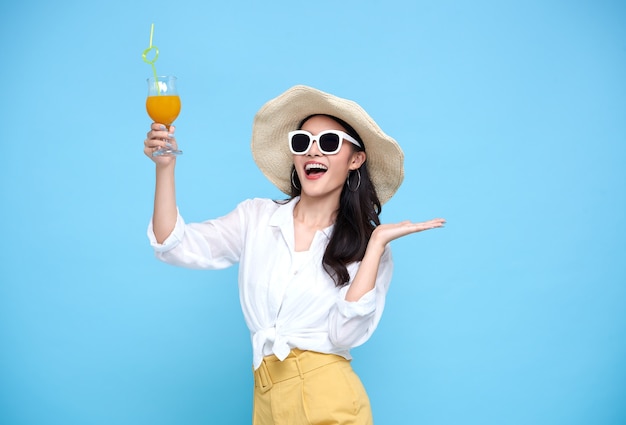 Glückliche asiatische Frau im Sommer lässige Kleidung, die Strohhut, Sonnenbrille hält Glas des frischen Fruchtsaftgetränks lokalisiert auf hellblauer Wand