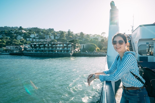 Glückliche asiatische Frau mit Sonnenbrille genießt die schöne Meereslandschaft auf einer Fähre mit Händen, die an einem sonnigen Tag in Kalifornien auf dem Geländer ruhen