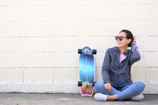 Glückliche asiatische Frau mit Sonnenbrille mit Surfskate-Skateboard, die gegen Betonwand sitzt