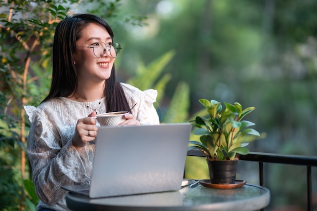 Glückliche asiatische freiberufliche Geschäftsfrauen halten eine Kaffeetasse, die beiläufig mit Laptop-Computer mit Notebook und Smartphone im Café arbeitet, Business Lifestyle-Kommunikationskonzept