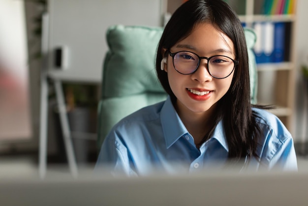 Glückliche asiatische Geschäftsfrau, die Computer trägt, die Ohrhörer tragen, die im Büro sitzen