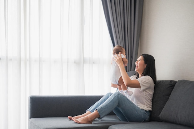 Glückliche asiatische lachende Mutter, die ihr entzückendes 3-Motten-Baby auf dem Sofa im Wohnzimmer in die Luft hebt