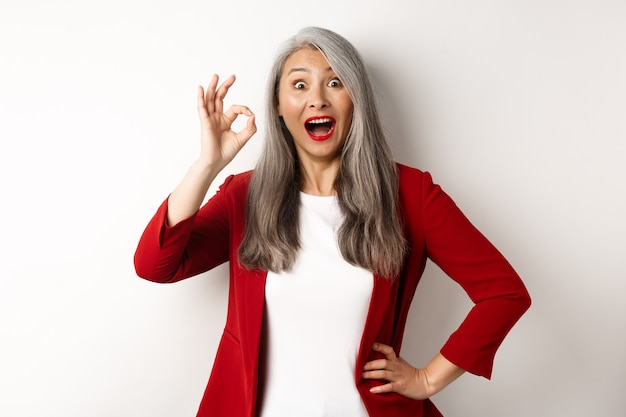 Glückliche asiatische reife Frau im roten Blazer, der okay Zeichen zeigt, erstaunt lächelt, etwas Fantastisches lobend, über weißem Hintergrund stehend.