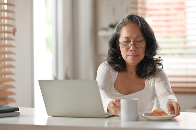 Glückliche asiatische Seniorin beim Frühstück und mit Laptop auf dem Schreibtisch zu Hause Älteres Lebensstil- und Technologiekonzept