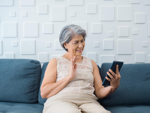 Glückliche asiatische Seniorin, die auf der Couch sitzt und im weißen Raum auf dem digitalen Tablet-Bildschirm in der Hand grüßt Ältere Frauen treffen sich mit der Familie per Videoanruf zu Hause Ältere Menschen mit Technologie