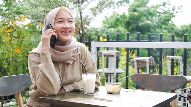 Glückliche Asien-Hijab-Frau, die Smartphone benutzt und anruft, im Café sitzt