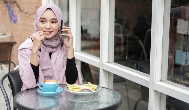 Glückliche Asien-Hijab-Frau, die Smartphone benutzt und anruft, Kaffee trinken, während im Café sitzen