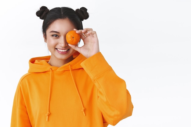 Glückliche attraktive asiatische Frau mit süßen Haarknoten, orangefarbenem Hoodie, eine Mandarine über dem Auge halten und lächelnde Kamera, Konzept eines gesunden Lebensstils, natürliches Make-up, weiße Wand stehen