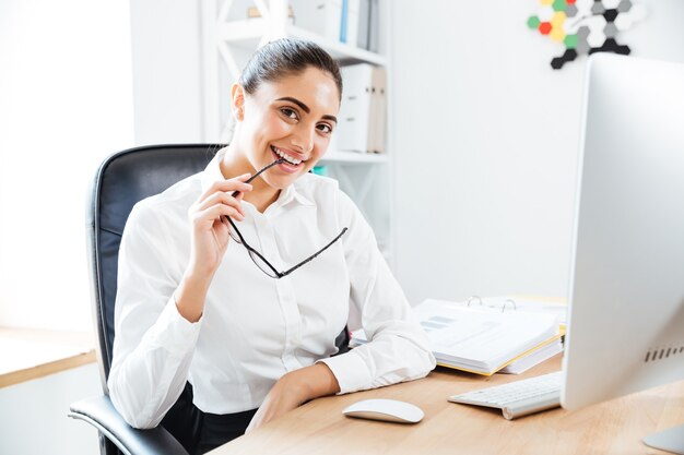 Glückliche attraktive Geschäftsfrau, die eine Brille hält und im Büro sitzt