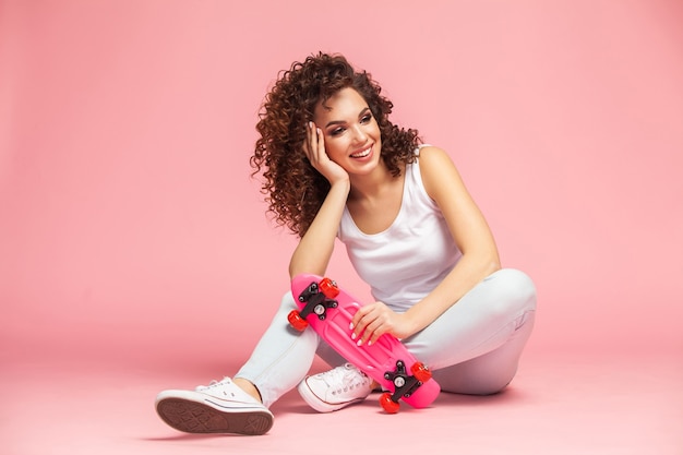 Glückliche attraktive junge Frau, die auf Skateboard über Rosa sitzt.