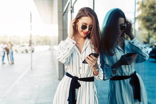 Glückliche attraktive junge Frau in der Sonnenbrille, die Smartphoneschirm betrachtet