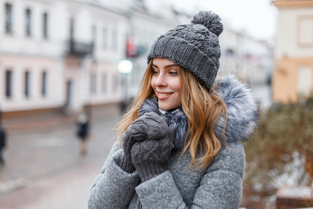 Glückliche attraktive junge Frau in einer Winterstrickmütze in einem trendigen grauen Mantel mit Fell in gestrickten warmen Handschuhen mit einem schönen Lächeln geht an einem Wintertag durch die Stadt. Fröhliches Mädchen genießen einen Spaziergang.