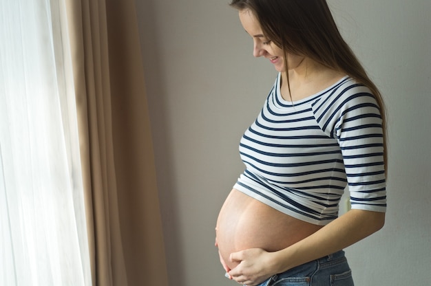 Glückliche attraktive schwangere Frau, die nahe am Fenster steht und ihren Bauch hält. Konzepte von Schwangerschaft und Familie