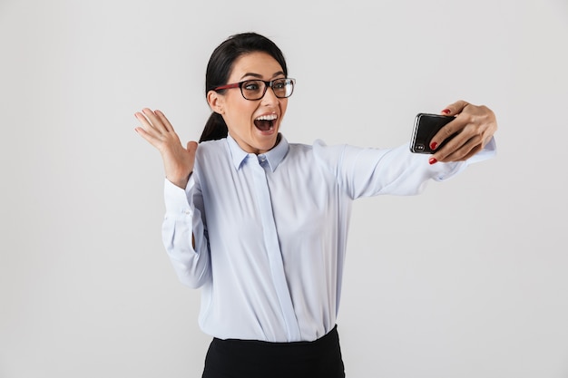Glückliche aufgeregte Geschäftsfrau, die klug gekleidet steht, isoliert über weißer Wand steht und ein Selfie nimmt