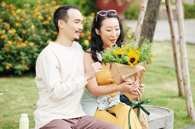 Glückliche aufgeregte junge Chinesin, die einen schönen Blumenstrauß von ihrem Freund betrachtet, wenn sie auf der Bank im Park sitzen?