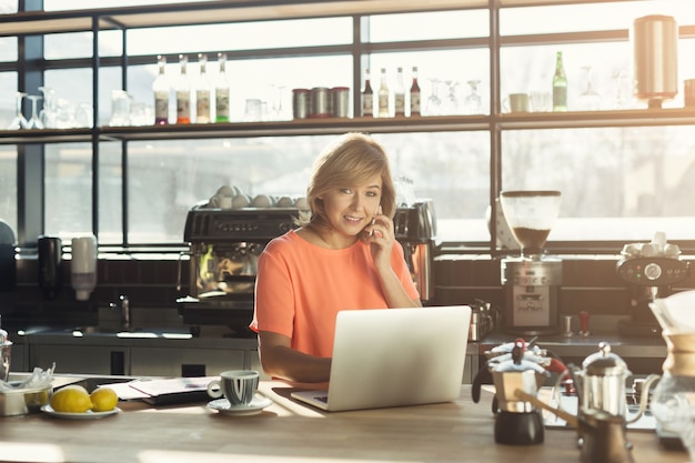 Glückliche Barista mittleren Alters, die am Laptop arbeitet und auf dem Smartphone spricht und eine Bestellung im modernen Café entgegennimmt. Kleinunternehmen, Online-Kauf- und Servicekonzept, Kopierraum