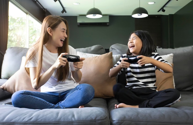 Glückliche Beziehung kleines Mädchen mit Mutter mit Joystick beim Spielen von Videospielen, die zusammen auf dem Sofa im Wohnzimmer sitzen und Familienurlaub genießen