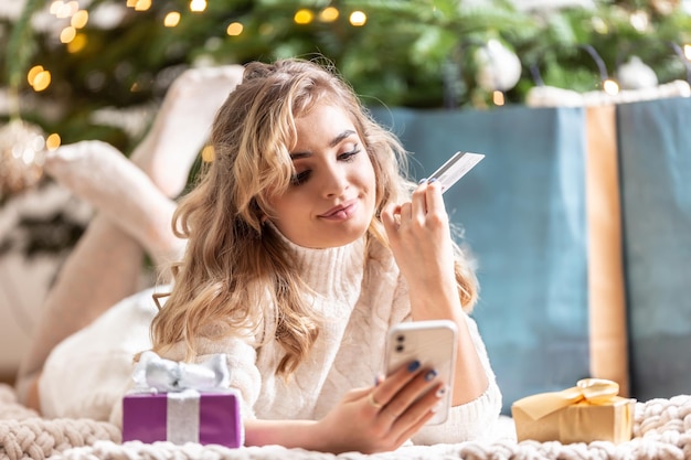 Glückliche blonde Frau, die Smartphone und Kreditkarte beim Online-Shopping verwendet, liegt zu Hause auf einer Decke in der Nähe von Geschenken und Weihnachtsbaum