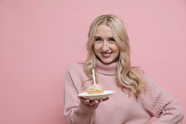 Glückliche blonde Frau im rosa Pullover, der Kuchen mit Kerzen hält und Geburtstag feiert. Speicherplatz kopieren