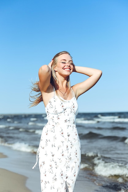 Glückliche blonde Frau in freier Glückseligkeit am Meeresstrand, die gerade steht. Porträt eines weiblichen Models im weißen Sommerkleid, das die Natur während des Reiseurlaubs im Freien genießt