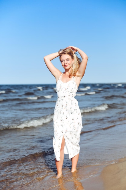 Glückliche blonde Frau in freier Glückseligkeit am Meeresstrand, die gerade steht. Porträt eines weiblichen Models im weißen Sommerkleid, das die Natur während des Reiseurlaubs im Freien genießt